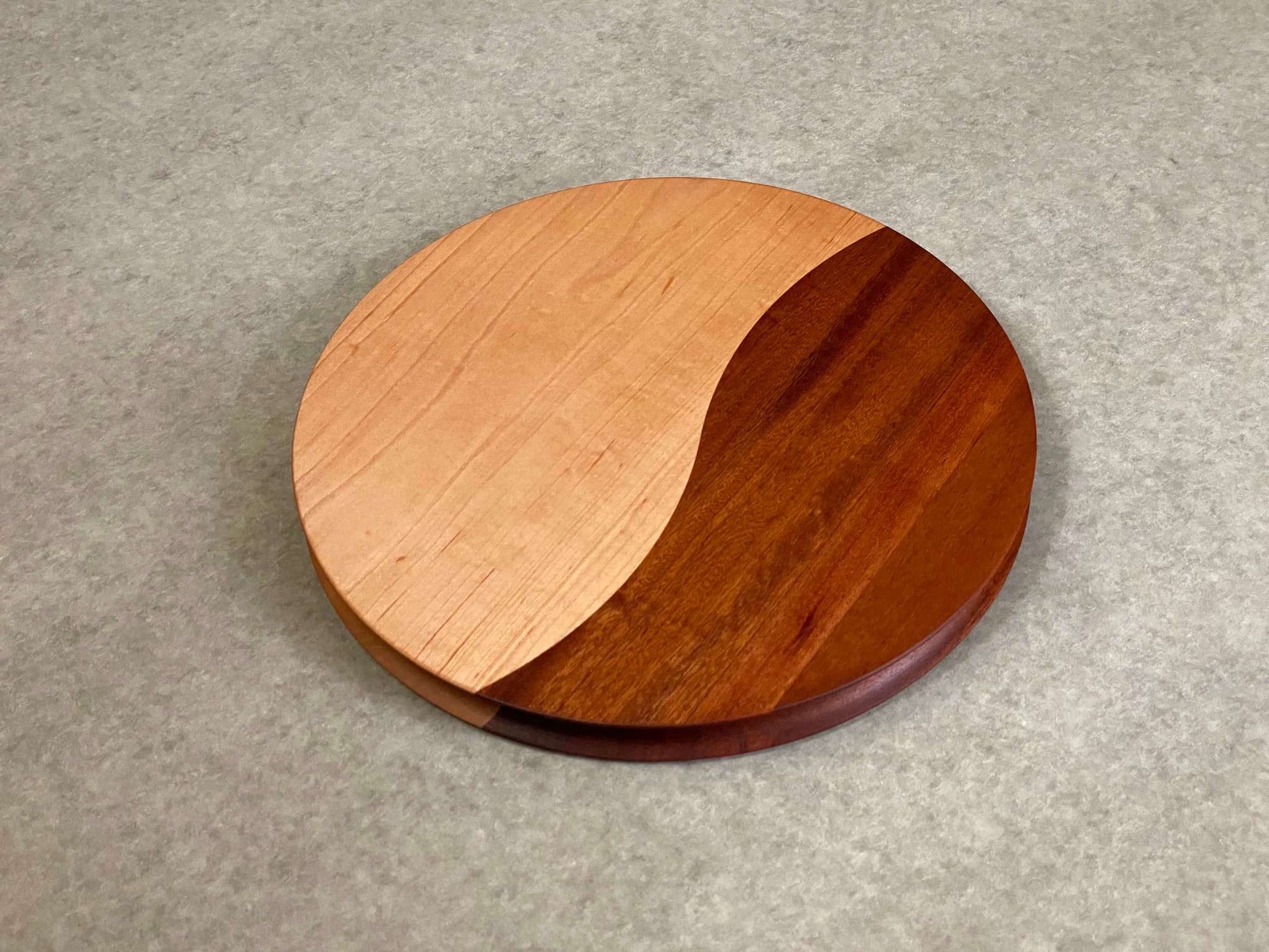 A round cutting and serving board made of part walnut and part mahogany with an off center curving joint. Sculpted edges provide a nice detail.
