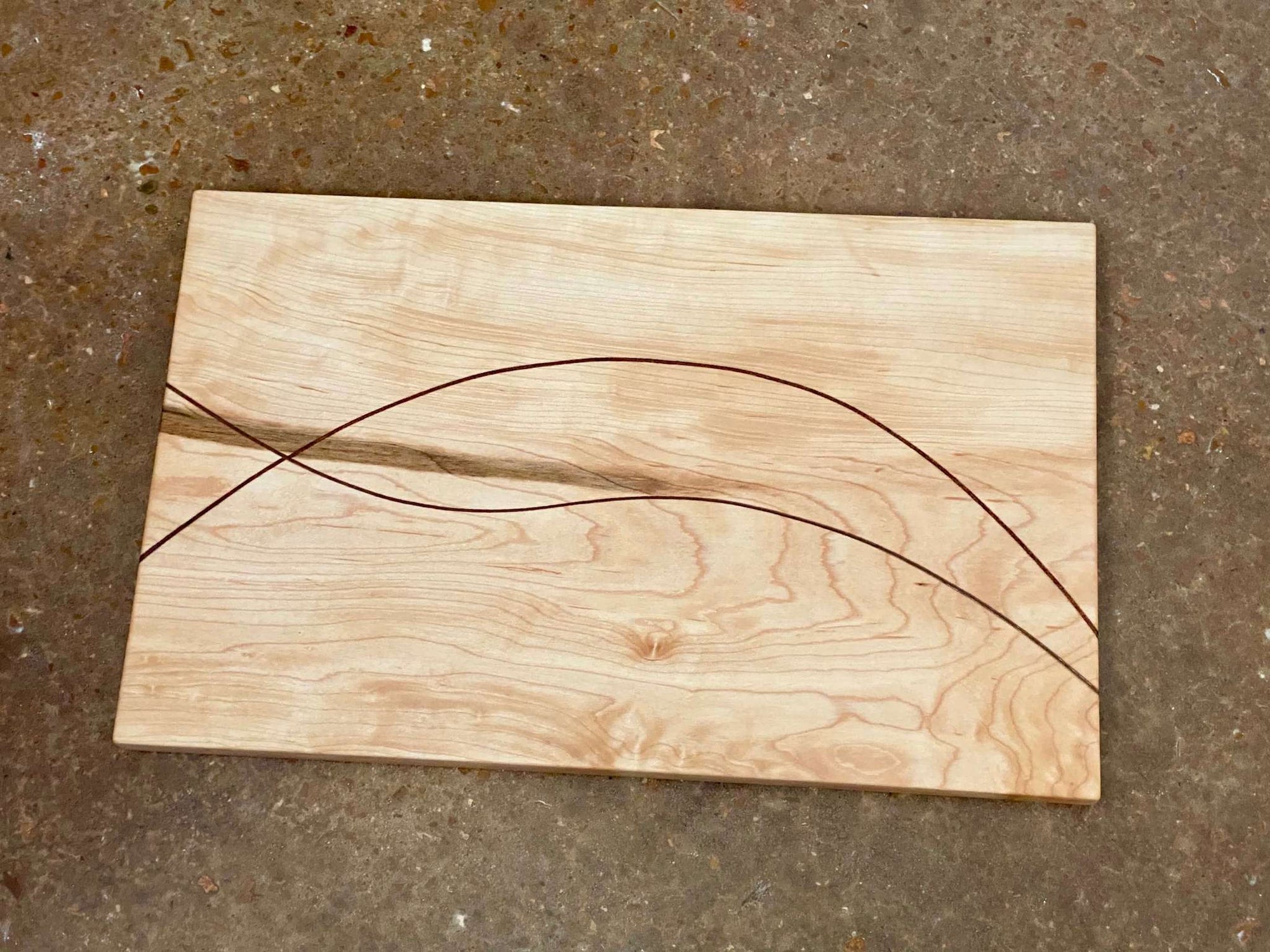 A rectangular cutting and serving board made of maple with two thin lines of walnut curving and intersecting through it.