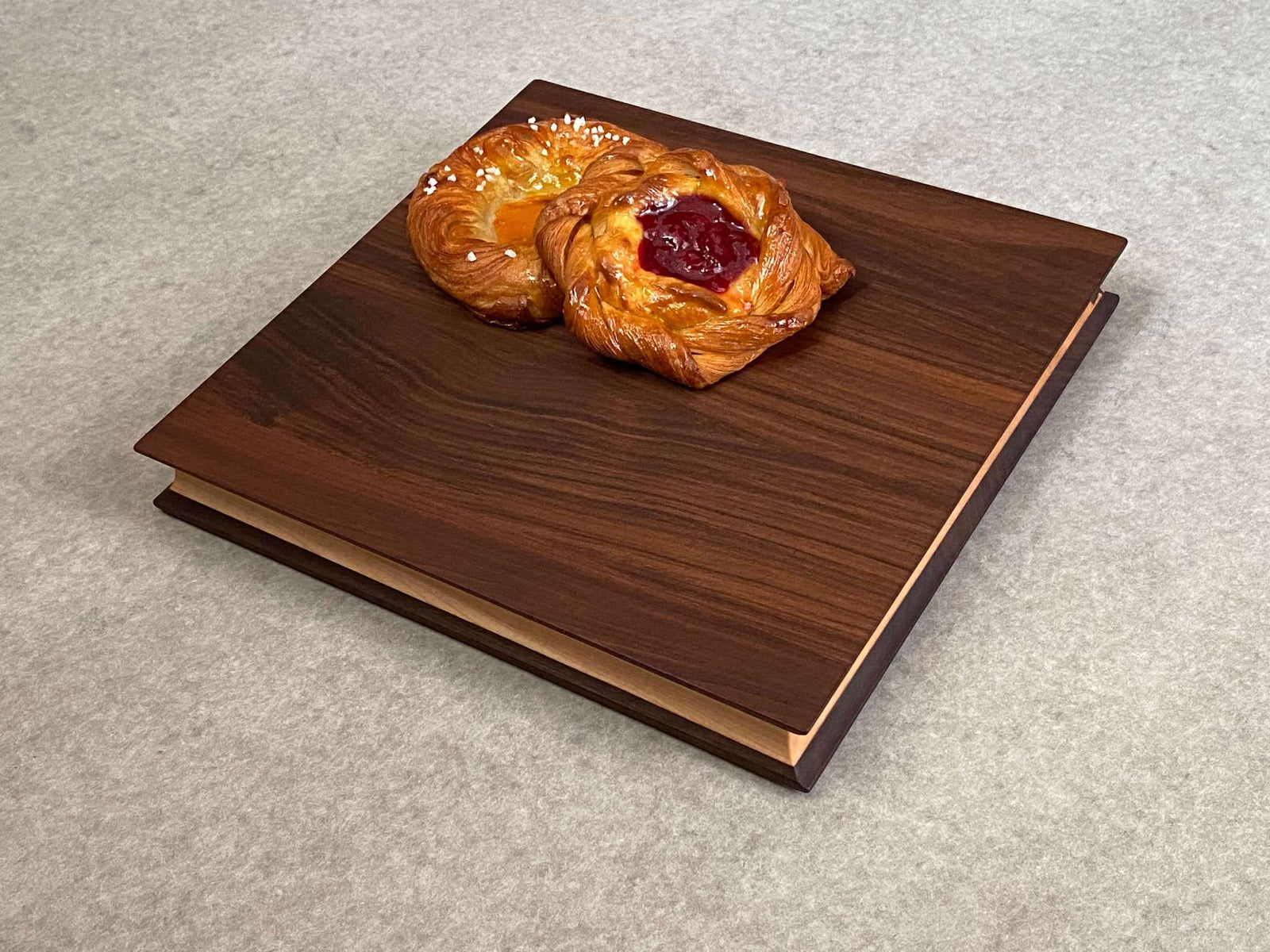 A square cutting and serving board of maple sandwiched between two thin layers of walnut. Sculpted edges provide comfortable fingerholds.