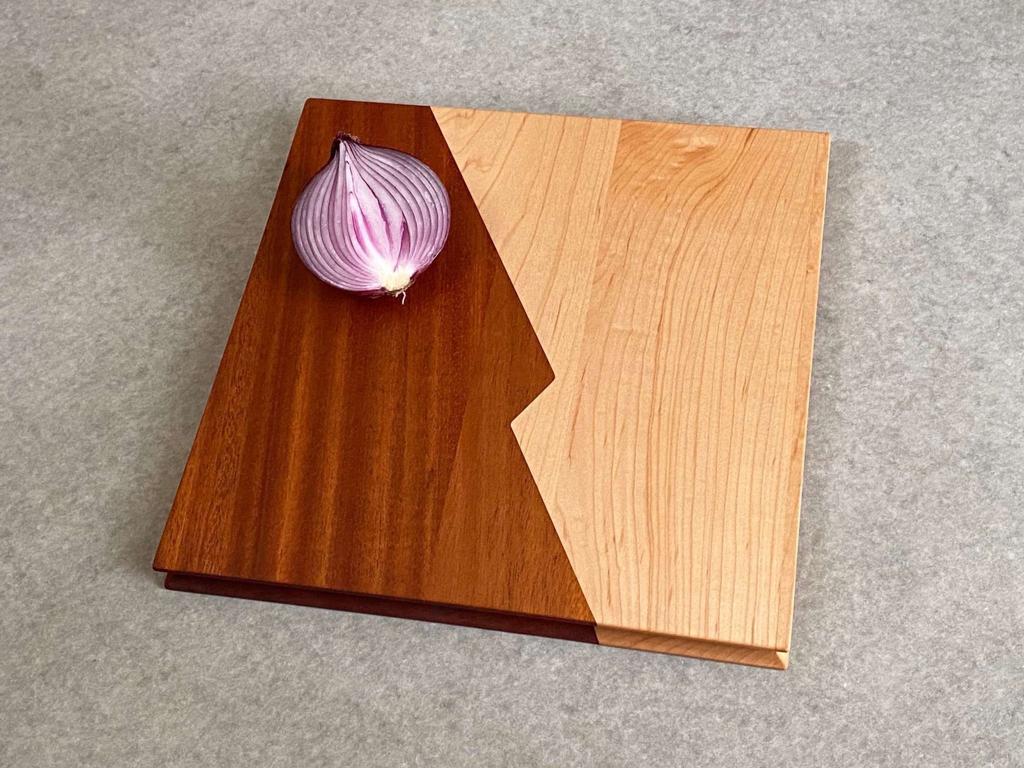 A square cutting and serving board that is half maple and half mahogany with a zigzag center joint. Sculpted edges provide comfortable fingerholds.